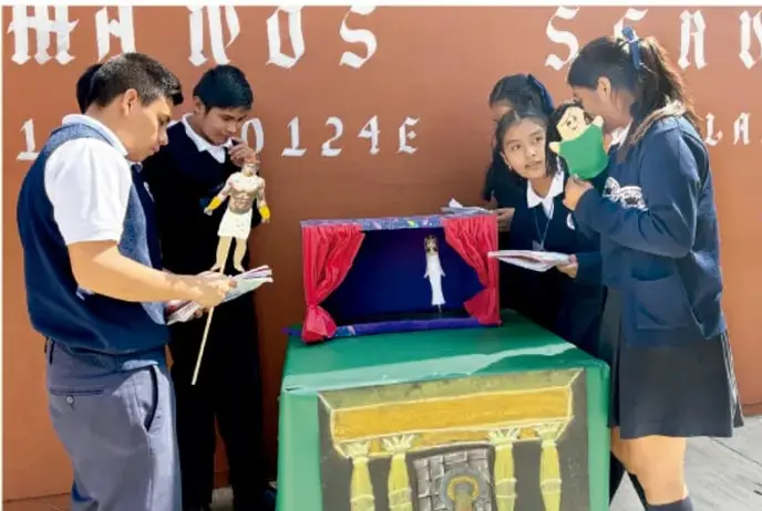 Fotografía de estudiantes de telesecundaria realizando una representación con títeres en un teatrito improvisado sobre una mesa, apoyada por una estructura pintada con columnas, mientras otros estudiantes observan con cuadernos.