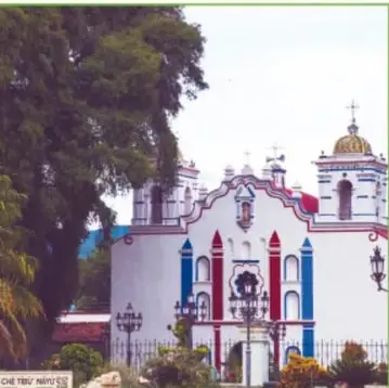 Fachada del templo de Santa María del Tule, blanco con adornos rojos y azules y portón con reja, frente a árboles.
