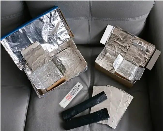 Dos cajas de cartón forradas con papel aluminio sobre un sillón, junto a un termómetro, tiras de cartón y tiras de papel aluminio, mostrando los materiales del experimento.
