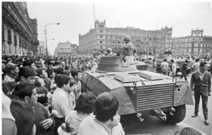 Fotografía histórica en blanco y negro del movimiento estudiantil de 1968 en la Ciudad de México: una multitud de estudiantes rodea un vehículo blindado (transporte blindado de la policía/militar) en una avenida céntrica, con edificios coloniales y comerciales al fondo, ilustrando la tensión entre manifestantes y fuerzas del orden.