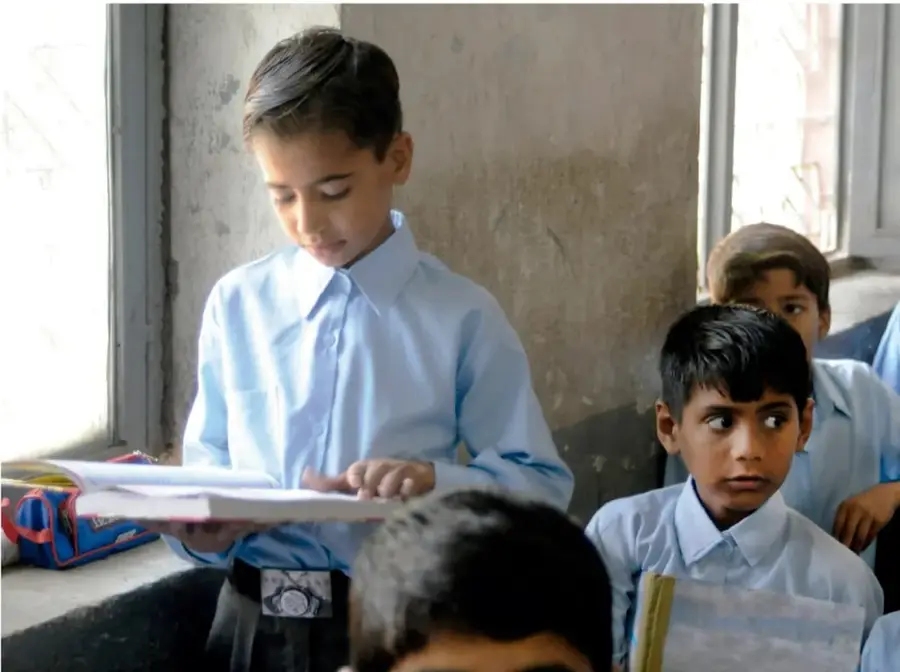 Fotografía de un niño con camisa azul leyendo un libro o cuaderno en un salón de clases, con otros compañeros alrededor.