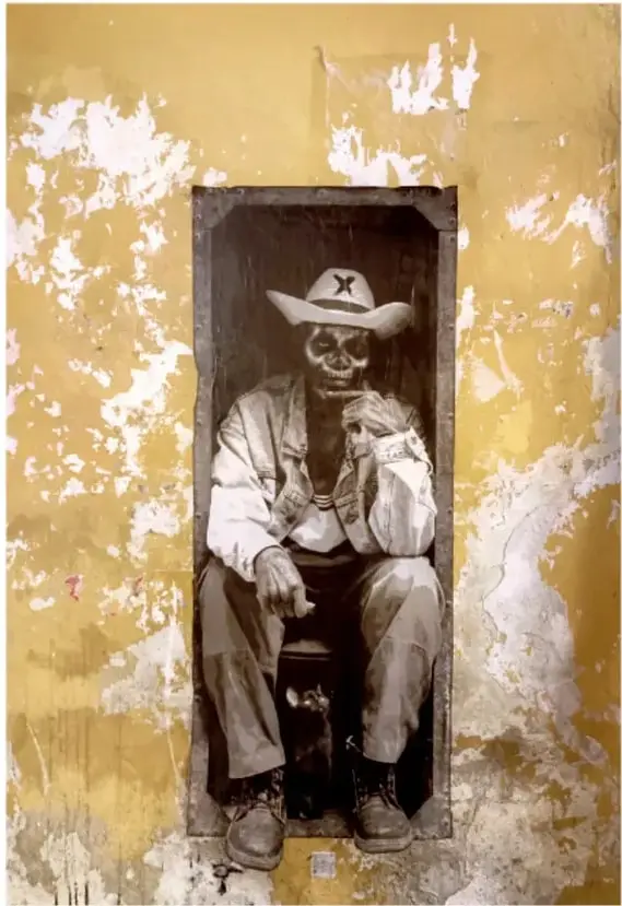Fotografía enmarcada: hombre con sombrero sentado en el vano de una ventana o puerta de un muro amarillo con pintura descascarada, en Oaxaca.