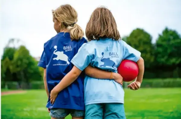 Fotografía de dos niñas de espaldas, tomadas de la cintura, con playeras azules que dicen 'Ball School'; una sostiene un balón rojo en un campo verde con árboles al fondo.