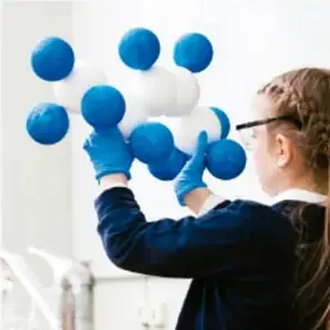 Estudiante con guantes manipulando un modelo molecular grande formado por esferas azules y blancas unidas, ilustrando la representación tridimensional de estructuras moleculares.