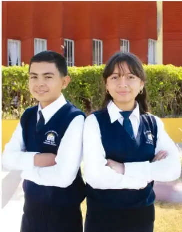 Fotografía de dos estudiantes de telesecundaria (un joven y una joven) con uniforme de chaleco azul y camisa blanca, de brazos cruzados, frente a un edificio y arbustos.