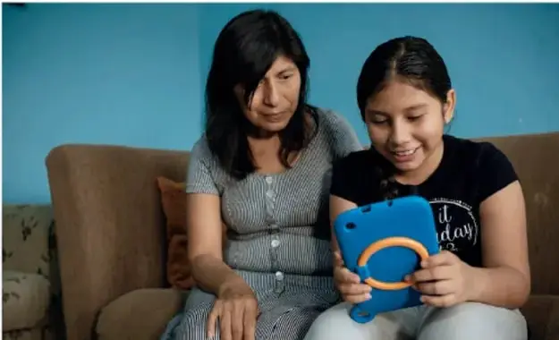 Fotografía de una mujer adulta y una niña sentadas en un sofá; la niña sostiene una tableta con funda azul y ambas observan la pantalla. Representa el uso de tecnología para informarse o comunicarse.