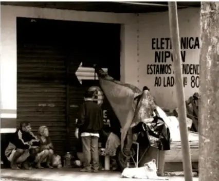 Personas en situación de calle sentadas frente a un local con letreros, en un entorno urbano.