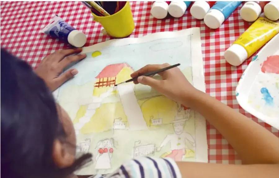 Fotografía de una niña pintando con acuarelas un dibujo sobre un mantel de cuadros rojos y blancos, rodeada de tubos de pintura y un vaso con pinceles.
