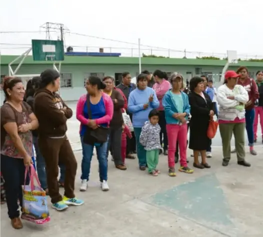 Fotografía de un grupo de personas, en su mayoría mujeres, reunidas en el patio de una escuela; al fondo se observa una cancha deportiva y edificios escolares.