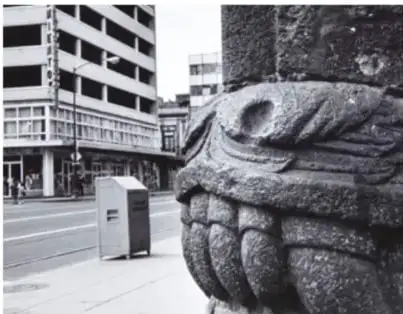 Fotografía de una escultura de piedra (cabeza de serpiente con dientes y ojos tallados) empotrada en la esquina de un edificio antiguo, con la calle y edificios modernos al fondo.