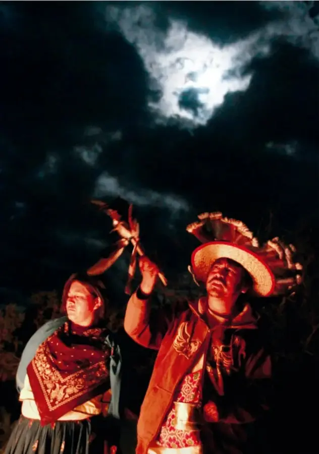 Fotografía nocturna de un mara'akame con sombrero tradicional y vestimenta bordada, con el brazo alzado y una antorcha, junto a otra persona; cielo nuboso iluminado por la luna.