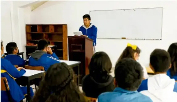 Fotografía de un estudiante de telesecundaria exponiendo de pie, con un libro en la mano, ante sus compañeros sentados en el aula.