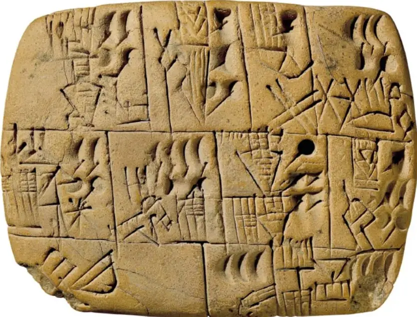 Tableta rectangular de barro color arena con muchas marcas talladas: cuñas, rayas, triángulos, círculos y figuras en casillas, semejante a una tablilla con escritura cuneiforme antigua.