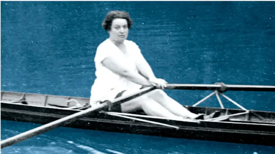 Fotografía histórica con tinte azul de una mujer con vestido blanco remando sola en una canoa o bote de remo sobre el agua, sosteniendo un remo de dos paletas.