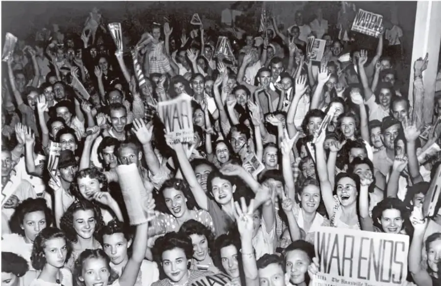 Fotografía en blanco y negro de una multitud celebrando el fin de la Segunda Guerra Mundial. Varias personas sostienen carteles con la frase 'WAR ENDS' (la guerra termina). Se observa alegría y euforia colectiva.