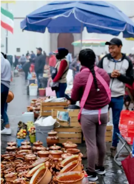 Fotografía de un mercado comunitario al aire libre con puestos de artesanías de barro, frutas y verduras; varias personas comprando y vendiendo bajo un toldo azul.