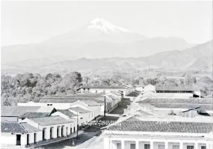 Fotografía antigua en blanco y negro de un pueblo con calles empedradas, casas de arcos y techos de teja, y un volcán nevado al fondo.