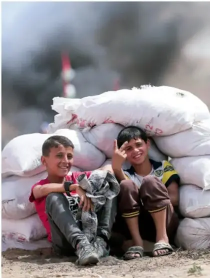 Fotografía de dos niños sonriendo sentados junto a costales de arena apilados como muro; la imagen remite a niños que viven contextos de conflicto y a la esperanza de la paz.