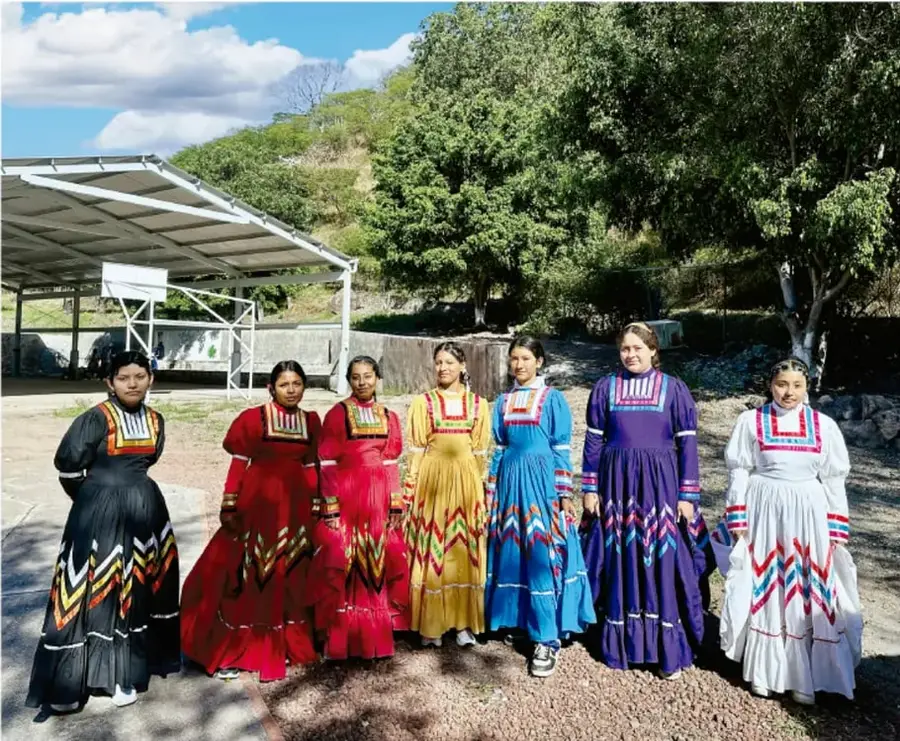 Fotografía de siete jóvenes indígenas mujeres con vestidos tradicionales de colores (negro, rojo, amarillo, azul, morado, blanco) con bordados geométricos, de pie al aire libre junto a una cancha techada y árboles, representando la diversidad de identidades juveniles en la comunidad.