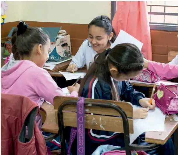 Fotografía de estudiantes de primaria trabajando en equipo en un salón de clases, sentados en pupitres, escribiendo y conversando.