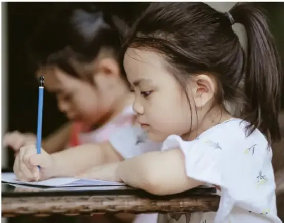 Fotografía de una niña escribiendo con un lápiz sobre una hoja, acompañada de otra compañera al fondo.