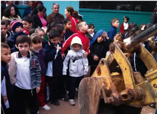 Estudiantes y adultos observan una excavadora amarilla en el patio de la escuela.