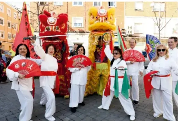 Fotografía de una celebración china en la calle: personas vestidas de blanco con abanicos rojos y trajes de león y dragón rojo y amarillo.