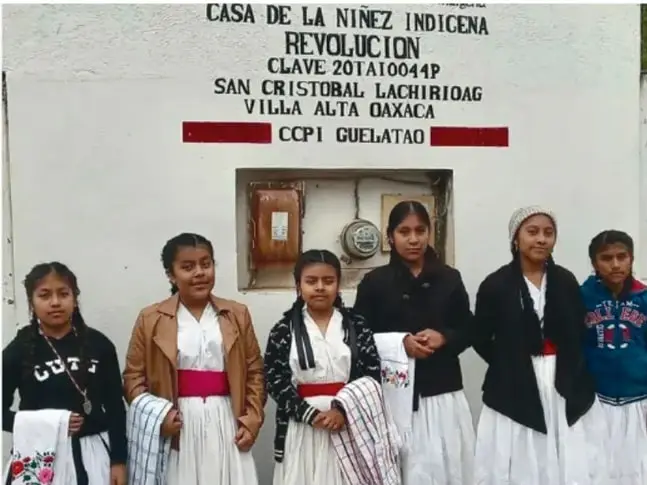 Seis jóvenes indígenas de pie frente a la fachada de la 'Casa de la Niñez Indígena Revolución' de San Cristóbal Lachirioag, Villa Alta, Oaxaca, con la leyenda CCP Guelatao.