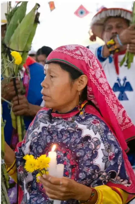 Fotografía de una mujer indígena con rebozo bordado y velas en las manos, junto a otras personas durante una ceremonia o ritual comunitario, en un espacio con adornos y plantas.