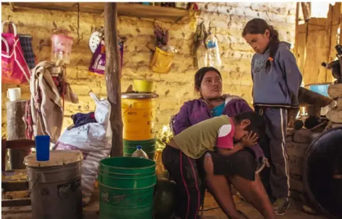 Fotografía de una familia en un espacio rural o precario; se observa a una mujer, una niña y un niño junto a utensilios y paredes de adobe, lo que ilustra condiciones de marginación y vivienda.