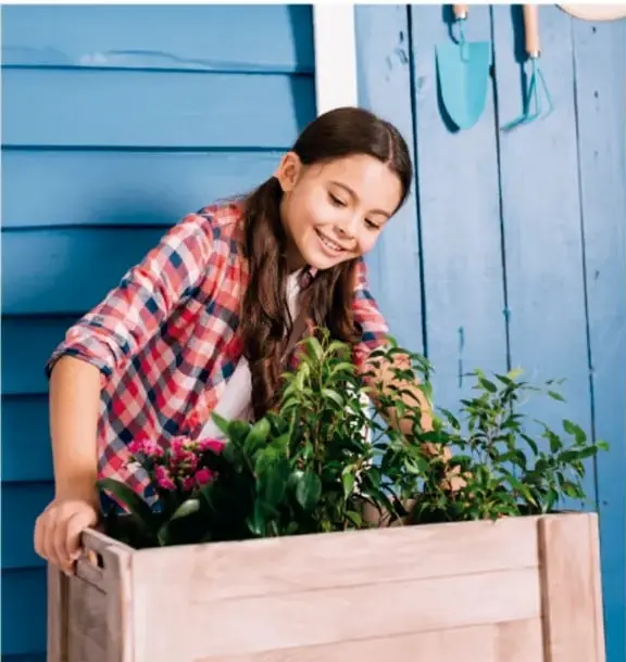 Fotografía de una niña sonriente con camisa de cuadros que sostiene un cajón de madera con plantas y flores, frente a una pared azul con herramientas de jardinería colgadas.