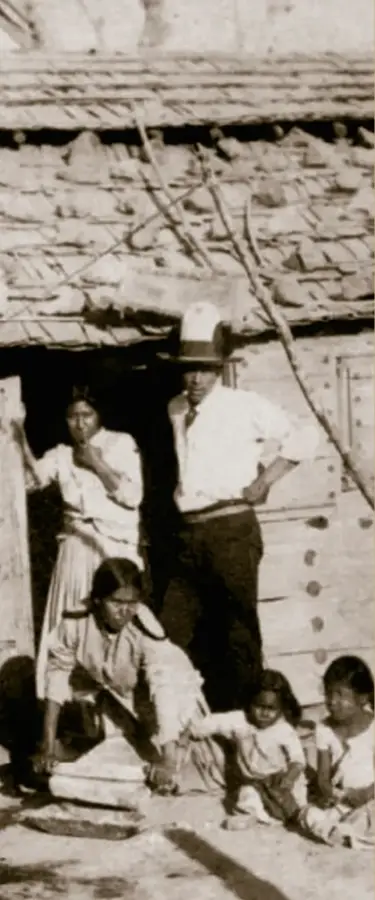 Fotografía sepia de una familia indígena frente a una choza de techo de teja; un hombre con sombrero y camisa blanca de pie, una mujer junto a él, otra mujer sentada en el suelo y dos niños pequeños en primer plano. Tarjeta de visita, ca. 1880. INAH/SINAFO.FN.MX.