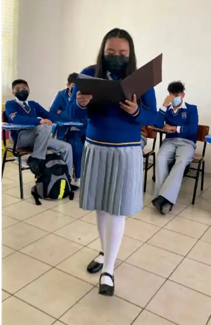 Fotografía de estudiantes en un salón de clases: una estudiante de uniforme azul y falda gris plisada lee en voz alta desde un libro, con cubrebocas; otros tres compañeros sentados en sus pupitres la escuchan, también con cubrebocas y uniforme escolar. Crédito: Luis Eduardo García Médel, INUXAI.