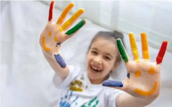 Niña riendo con fuerza, tapándose la cara con las manos, hombros encogidos.