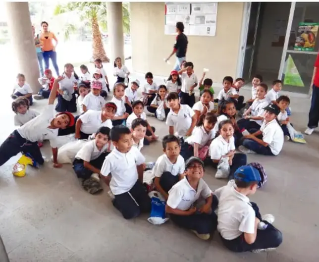 Fotografía de estudiantes de primaria con uniforme sentados en el suelo de un patio escolar, acompañados por una persona adulta; al fondo hay un edificio con carteles.