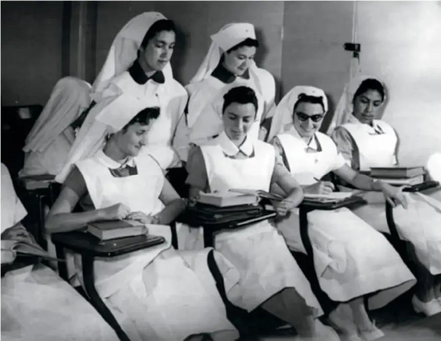 Fotografía en blanco y negro de un grupo de enfermeras o estudiantes de medicina con uniforme blanco y cofia, sentadas tomando apuntes en un aula.