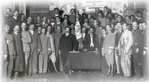 Fotografía histórica en blanco y negro de un grupo numeroso de personas, en su mayoría hombres con traje y sombrero, reunidas en un salón; al centro hay una mesa con documentos. Parece corresponder a una reunión o convención revolucionaria.