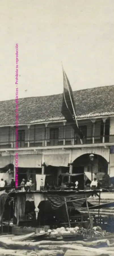 Fotografía histórica en blanco y negro de un puerto costero, con embarcaciones, personas y una edificación junto al agua.