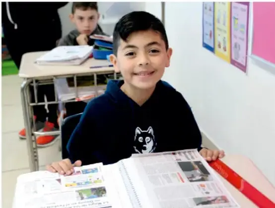 Fotografía de un niño sonriente sentado en su banca escolar, sosteniendo un periódico abierto; detrás se ve otro estudiante y un salón de clases con carteles.