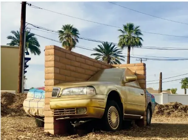 Obra *Amurallado* (2017): un auto color dorado incrustado en una pared de ladrillos que lo atraviesa, al aire libre con palmeras y cielo azul.