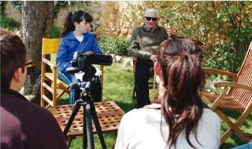 Fotografía de un grupo de estudiantes que entrevista a un señor mayor al aire libre; hay una cámara con trípode y sillas de madera, lo que ilustra el diálogo intergeneracional.