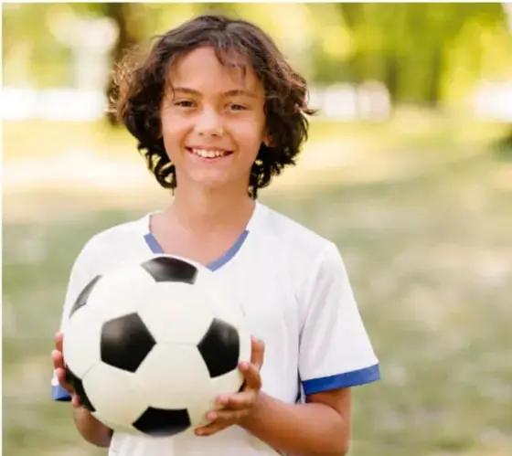 Niña sonriente con camiseta deportiva blanca sosteniendo un balón de fútbol, ilustrando la vida activa.