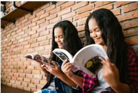 Foto de dos estudiantes sentadas frente a una pared de ladrillo, leyendo un libro juntas y sonriendo, como consulta de fuentes.