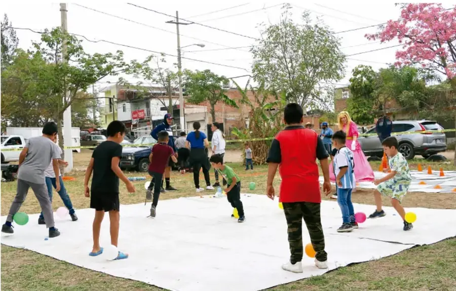 Fotografía de un grupo de niños y jóvenes en actividad recreativa al aire libre sobre una lona blanca extendida en el pasto, con balones de colores (blanco, verde, rosa, amarillo, naranja) y conos naranjas; al fondo hay árboles (uno con flores rosas), casas, autos y una persona disfrazada de princesa, sugiriendo una jornada comunitaria o recreativa.