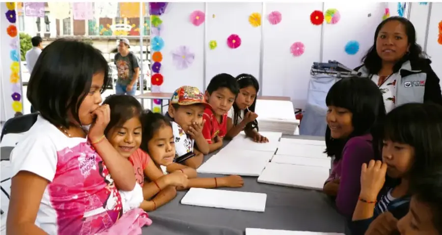 Fotografía de una asamblea escolar: niñas y niños sentados alrededor de una mesa con hojas blancas, una maestra de pie al fondo, decoración con flores de papel de colores en la pared.