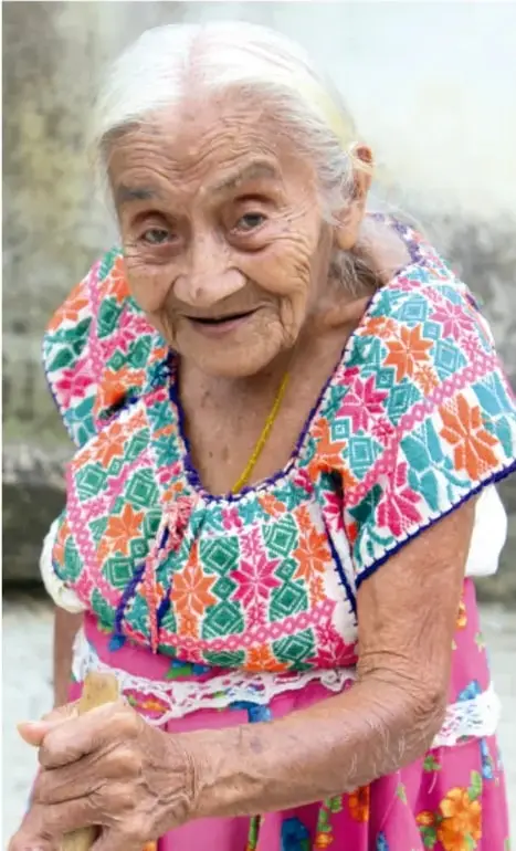 Fotografía de una mujer anciana indígena sonriente, con cabello blanco recogido, blusa bordada de colores vivos (rosa, verde, naranja) y falda rosa, apoyada en un bastón; representa a los mayores de la comunidad, tema relacionado con los roles según la edad.