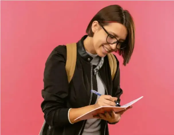 Fotografía de una joven estudiante con lentes, chamarra negra y mochila, escribiendo en un cuaderno sobre fondo rosa.