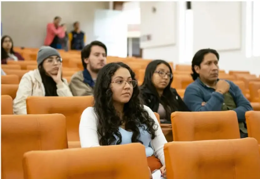 Fotografía de estudiantes universitarios o de secundaria sentados en un aula con butacas naranjas, atentos a una presentación o clase.