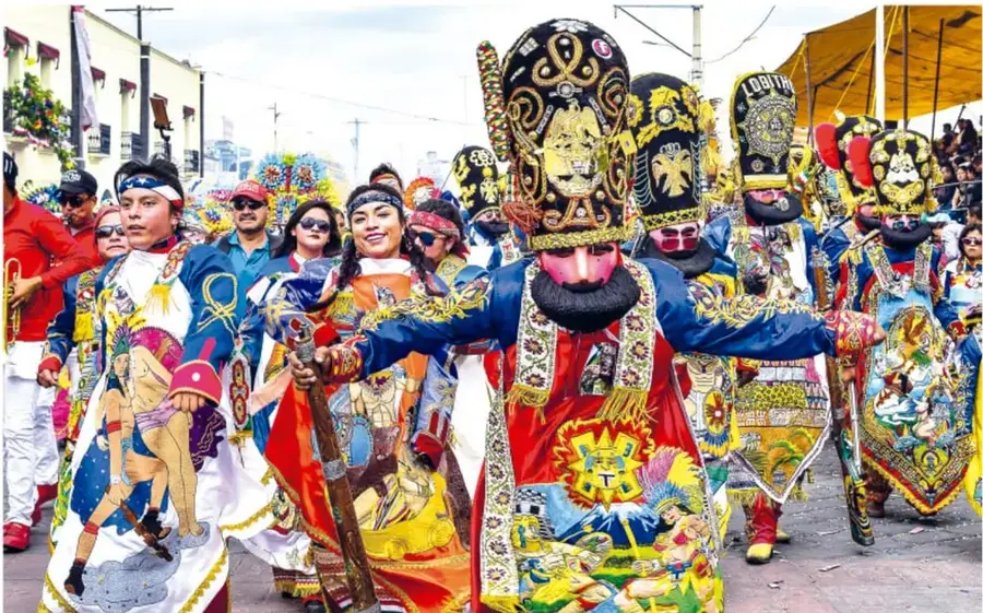 Desfile de carnaval mexicano: bailarines con penachos de plumas, máscaras con barba, trajes bordados coloridos y un personaje con bastón al centro.