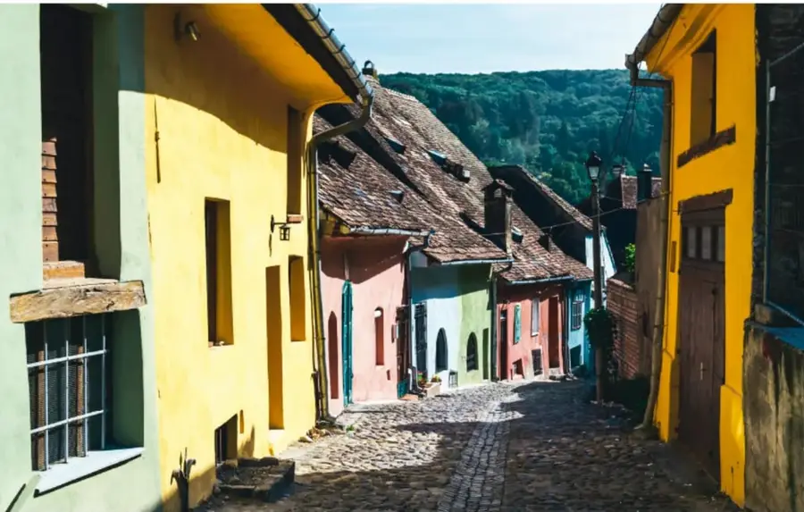 Calle empedrada con casas de colores amarillo, verde, rosa y azul, rodeada de cerros verdes; representa un paisaje típico de una comunidad.